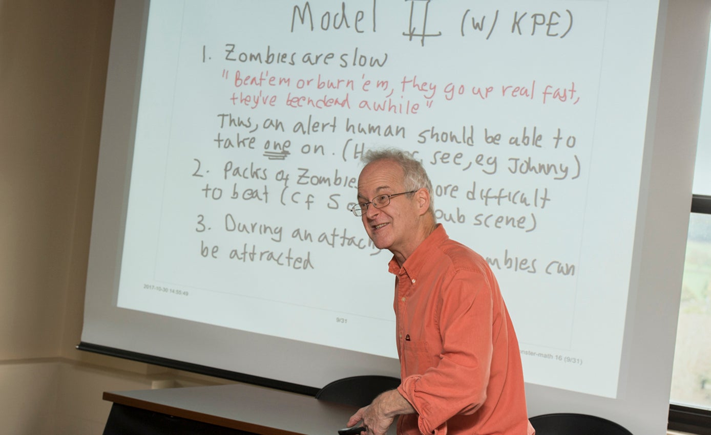 Ermentrout in an orange shirt in front of a screen with some rules of the math lesson on it