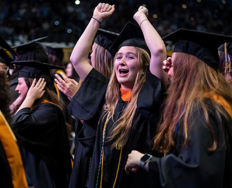 A person in a black graduation cap and gown celebrates with their hands up