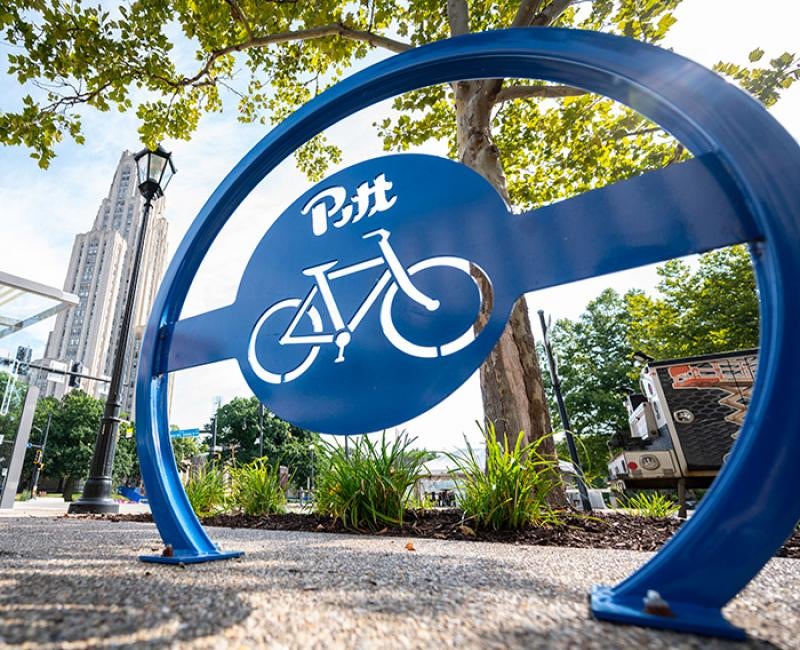 A blue bike stand with a cutout Pitt logo and bike in front of the Cathedral of Learning
