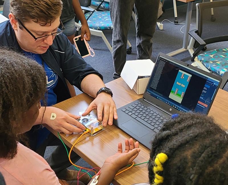 A teacher shows two students a control panel attached to a laptop