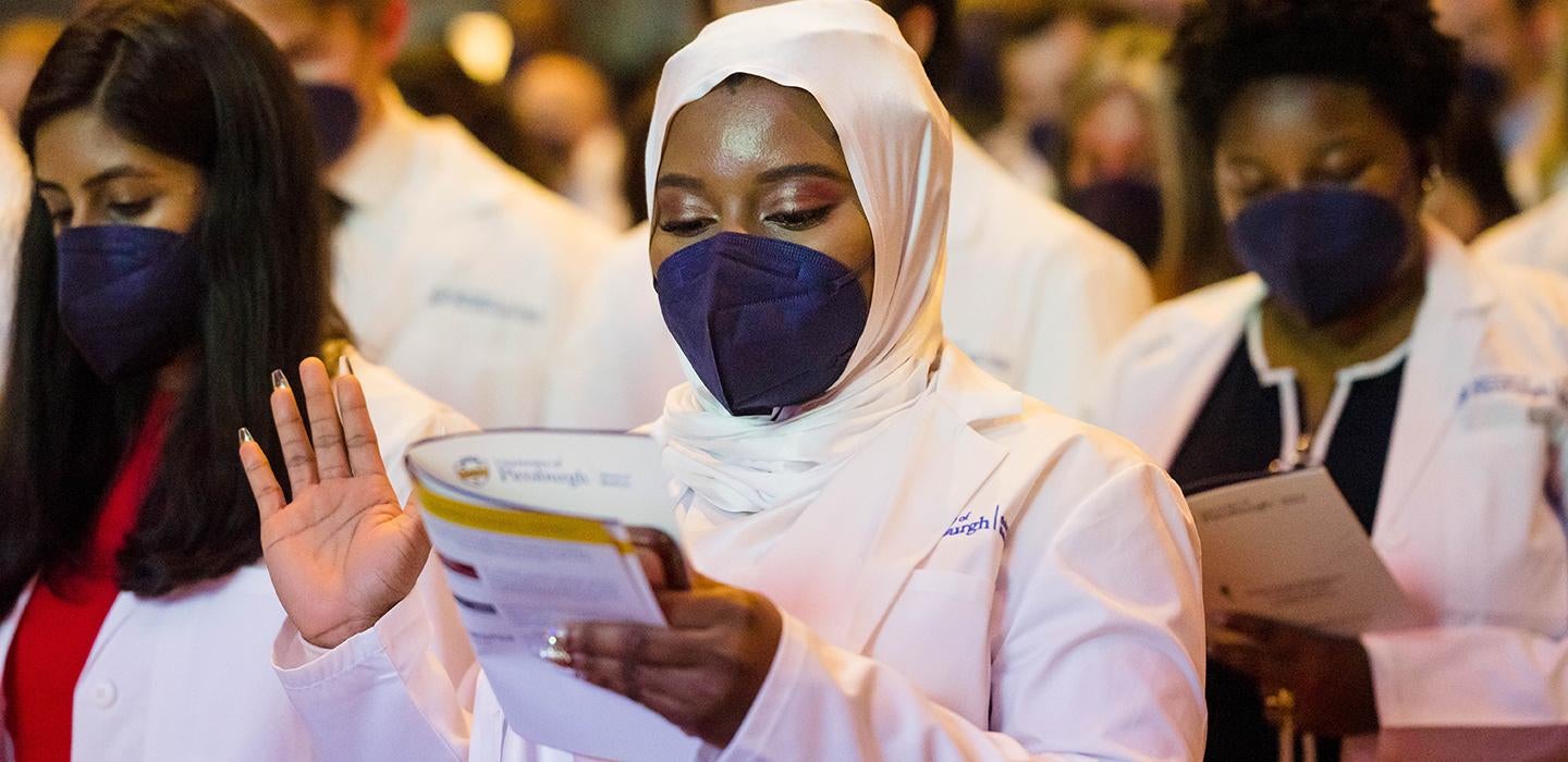 a woman in a blue mask, white coat and white head scarf holding up her right hand