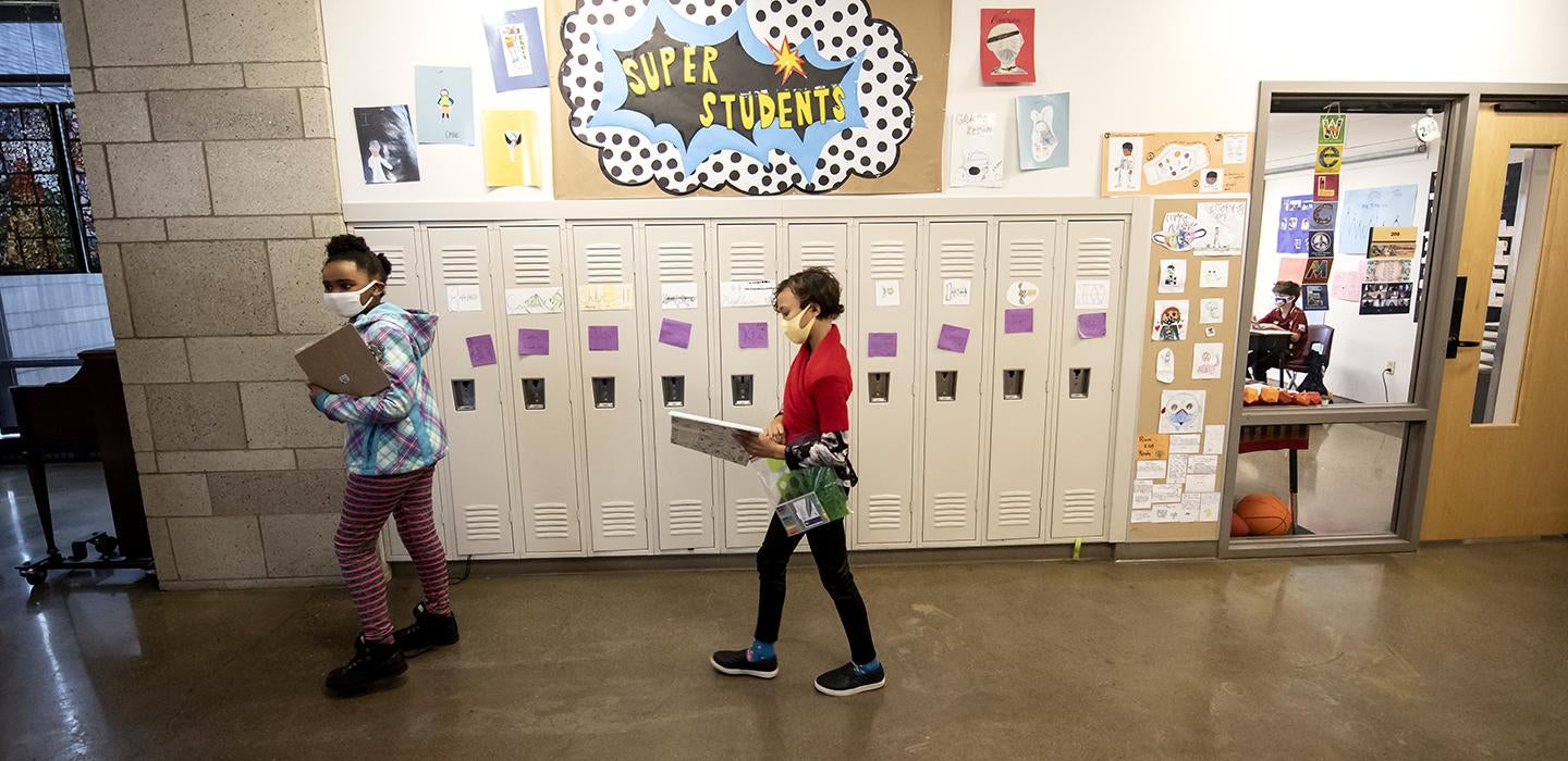Two students with masks walking through a hallway with lockers, a poster with "Super Students" hangs on the wall.