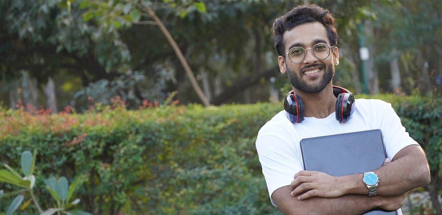 Student in white shirt holding laptop in front of shrubbery