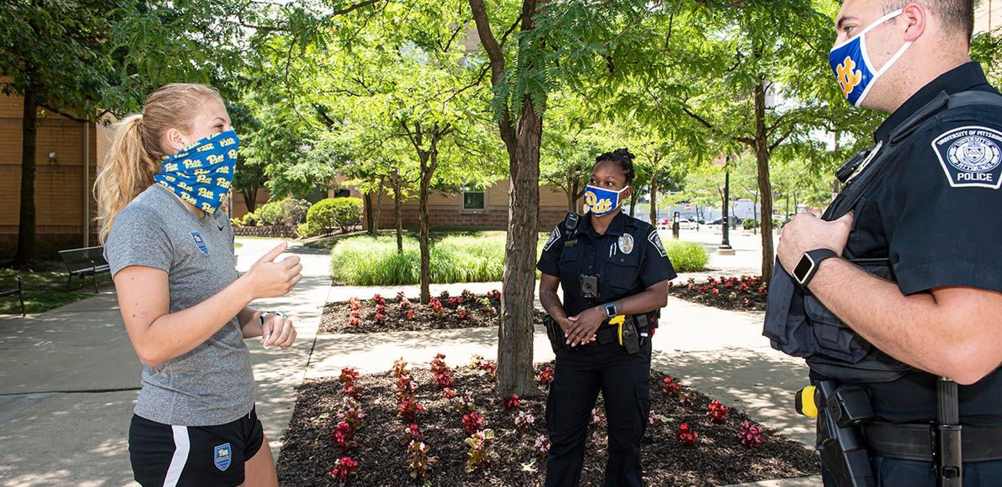 Two police officers in face masks talk to a person in a gray t-shirt and shorts