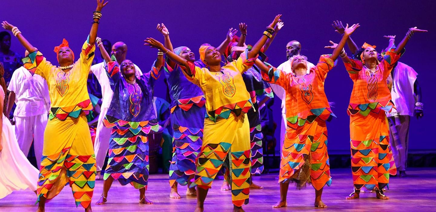 The KanKouran West African Dance Company dancing on stage in bright yellow and orange attire. 