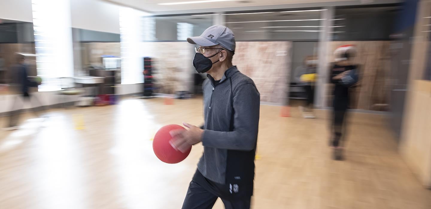 Man with grey shirt and hat holding a red ball in exercise room 