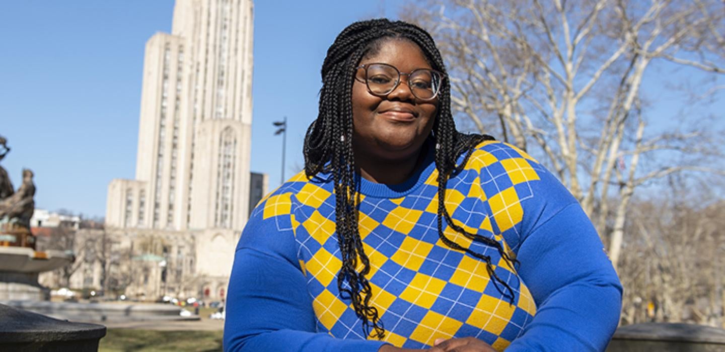 Haliyat Oshodi wearing blue and gold sweater posing in front of the Cathedral of Learning