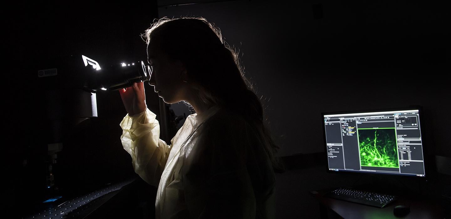 Woman looking through microscope in a dark room with computer in background