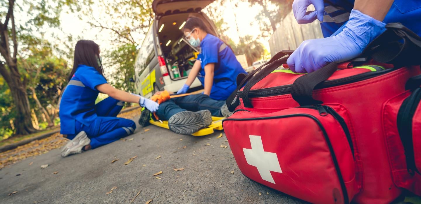 A gloved hand holds a red bag with a white cross as two people in scrubs help a person on the ground.