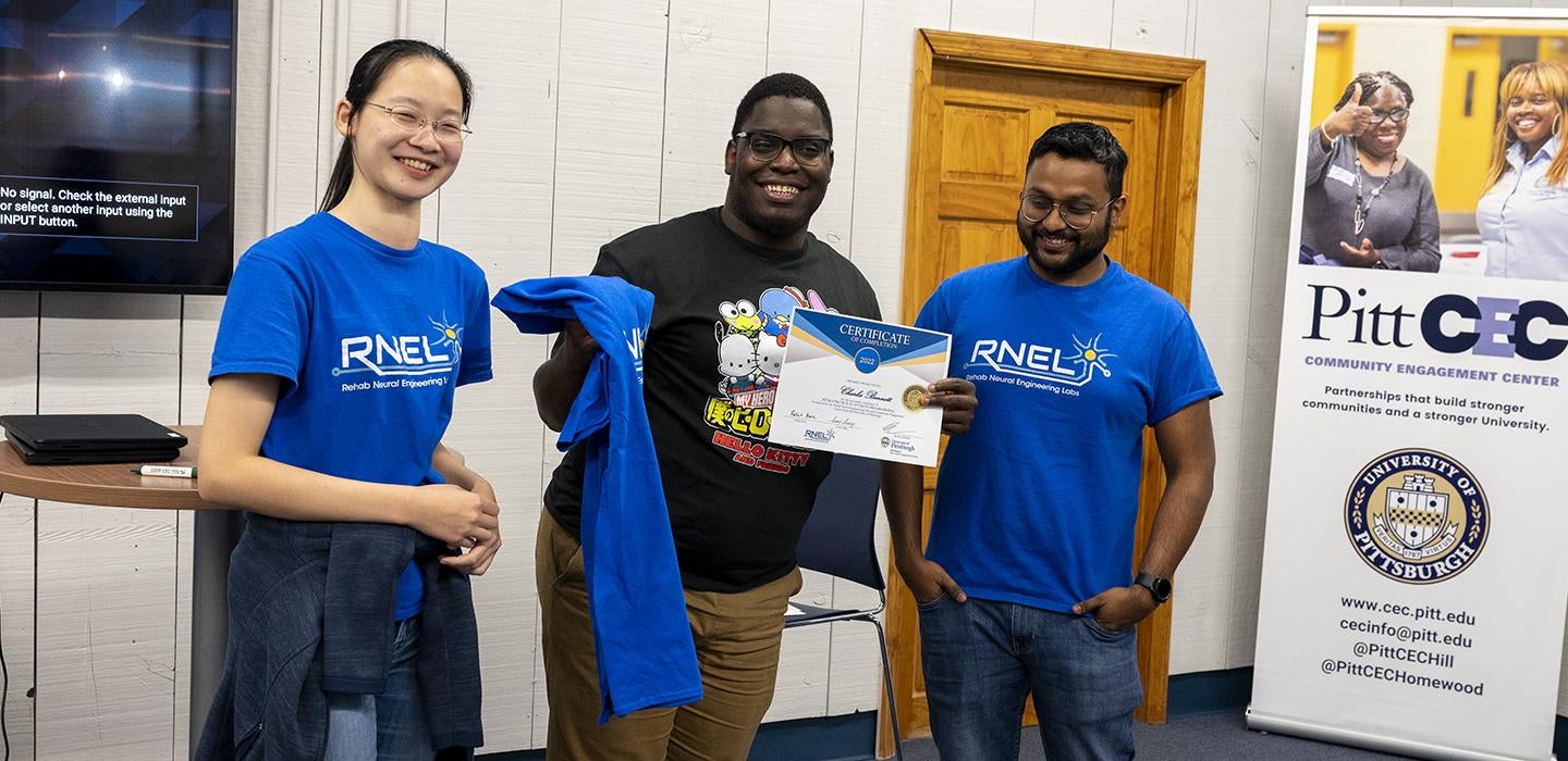 A person holds a certificate and T-shirt while standing in between two people in the same blue T-shirt that reads RNEL