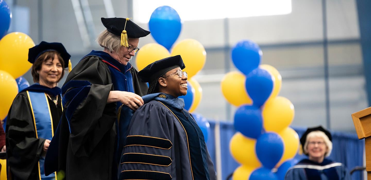 A person in doctoral regalia is hooded in front of blue and yellow balloons