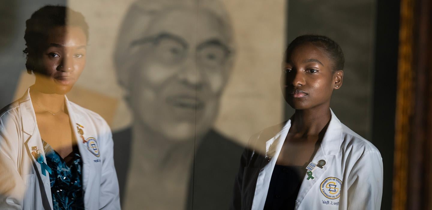 Two people in white coats stand in front of a portrait of Ella P Stewart