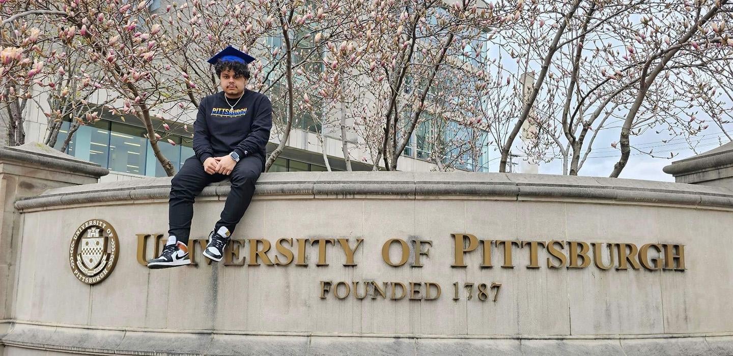 Knight sits on the marble University of Pittsburgh sign wearing all black and a blue graduation cap