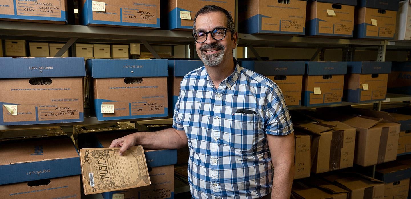 Cassaro holds an antique music book in front of a shelf of boxes of archives