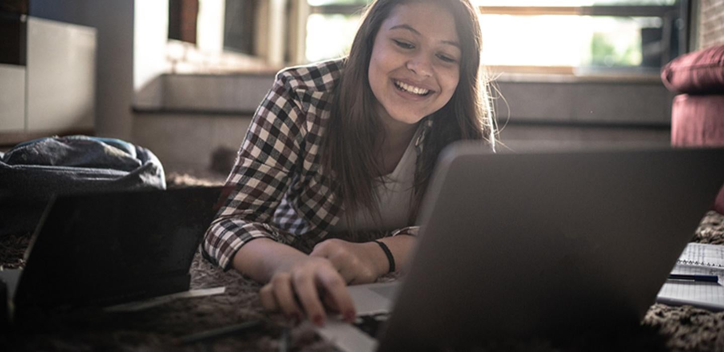 A girl lays on the floor, smiling into her laptop
