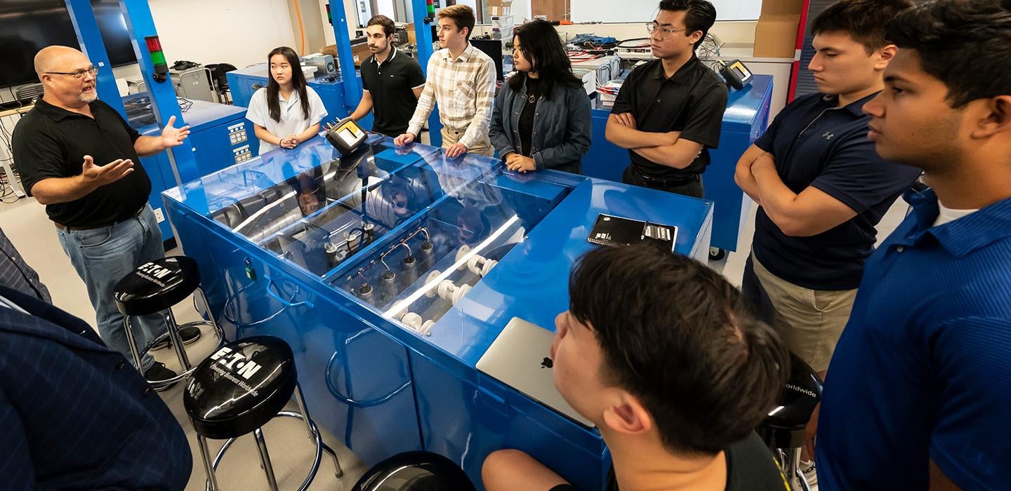 Students gather around a blue clear-top table in a lab and listen to an instructor