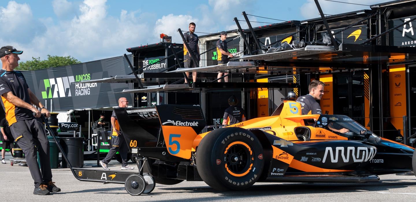 Team members pull an orange and black racing car from its garage onto the track