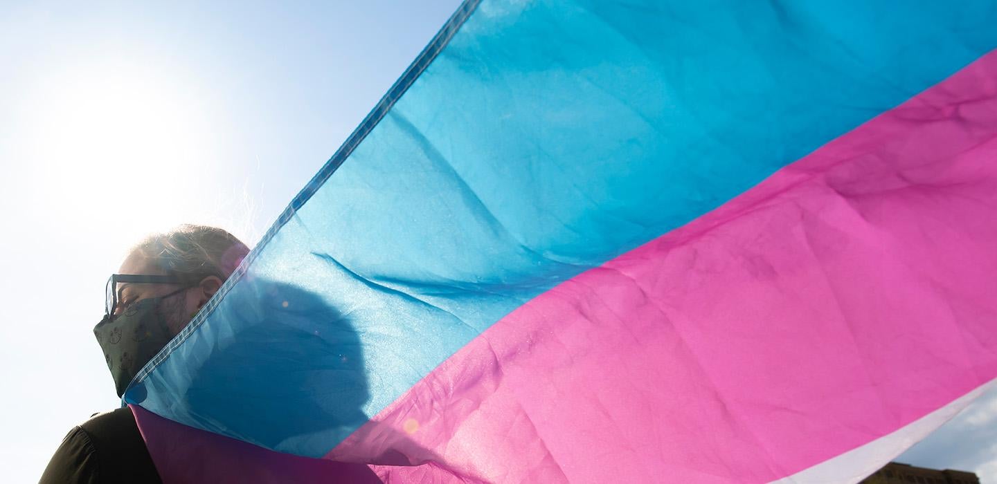 A person in a mask lets a transgender pride flag wave behind them
