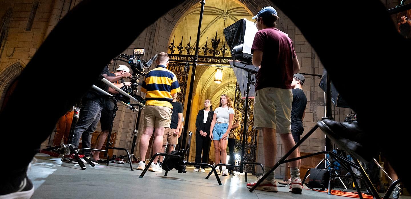A crew with cameras and lighting equipment films actors in the Cathedral of Learning
