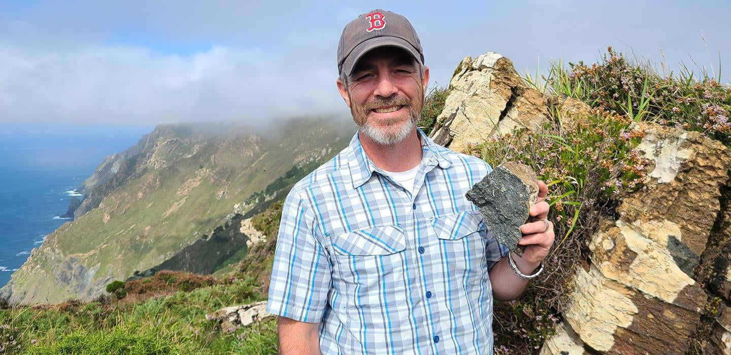 Kerrigan holds up a rock. A foggy seaside cliff is behind him.