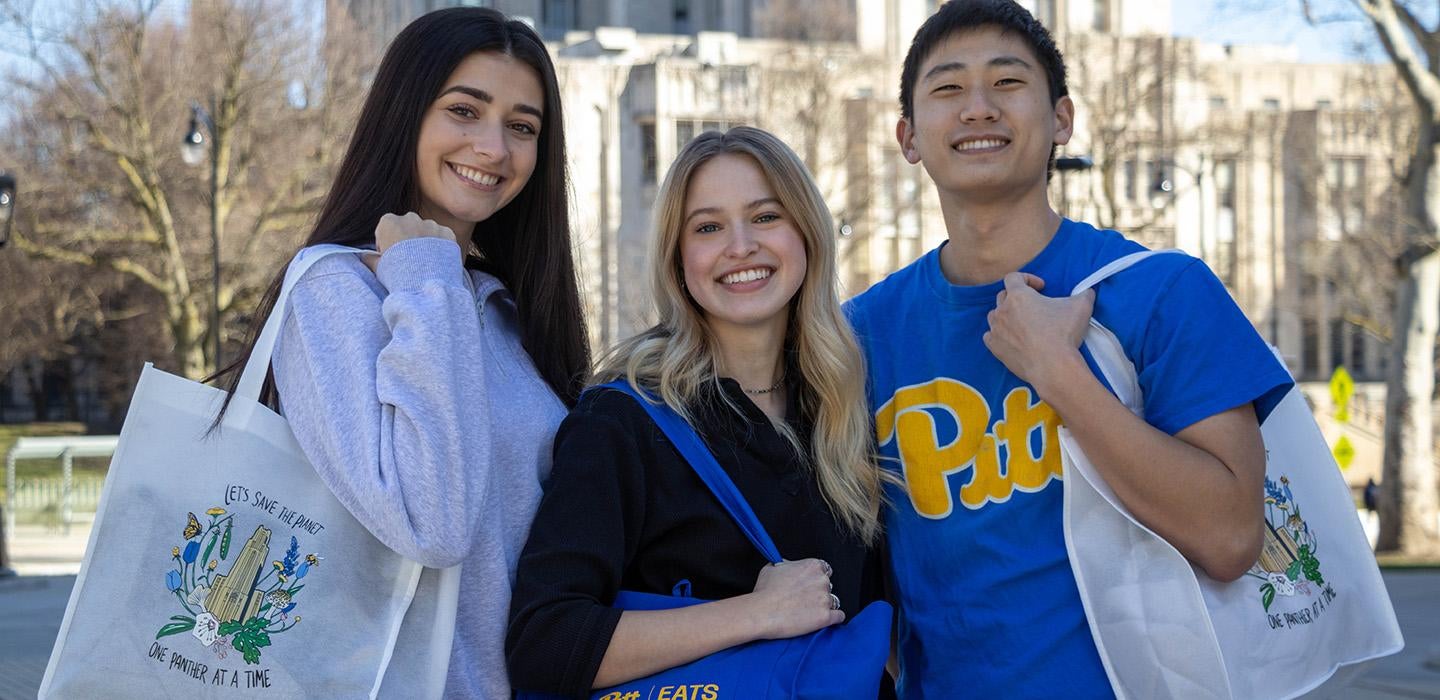 Three students in Pitt gear hold reusable grocery bags on their shoulders