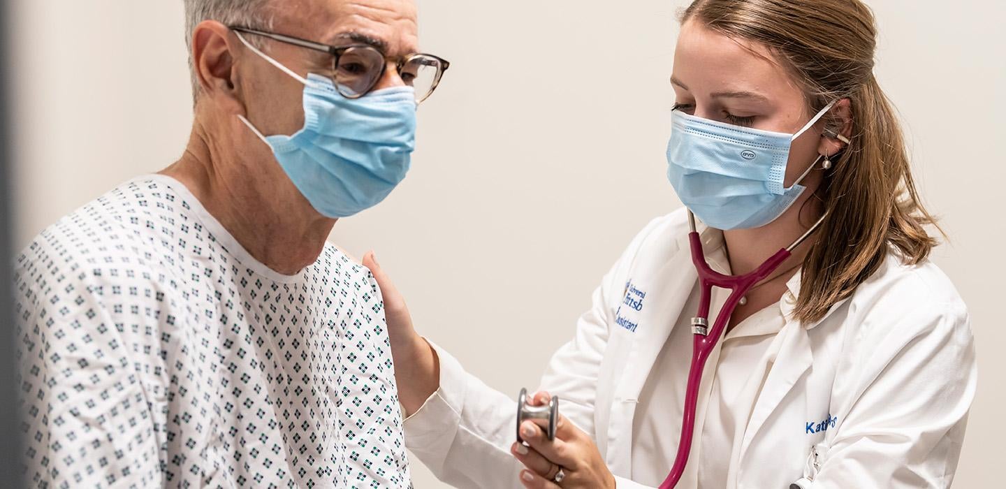 A doctor brings a stethoscope to a patient's chest. Both are wearing face masks