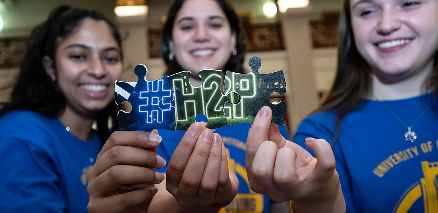 People hold up puzzle pieces to show a neon light that reads (hashtag) H 2 P