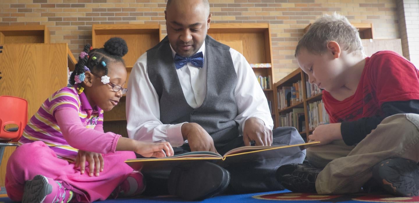 Male graduate student sitting on floor reading to two elementary school students