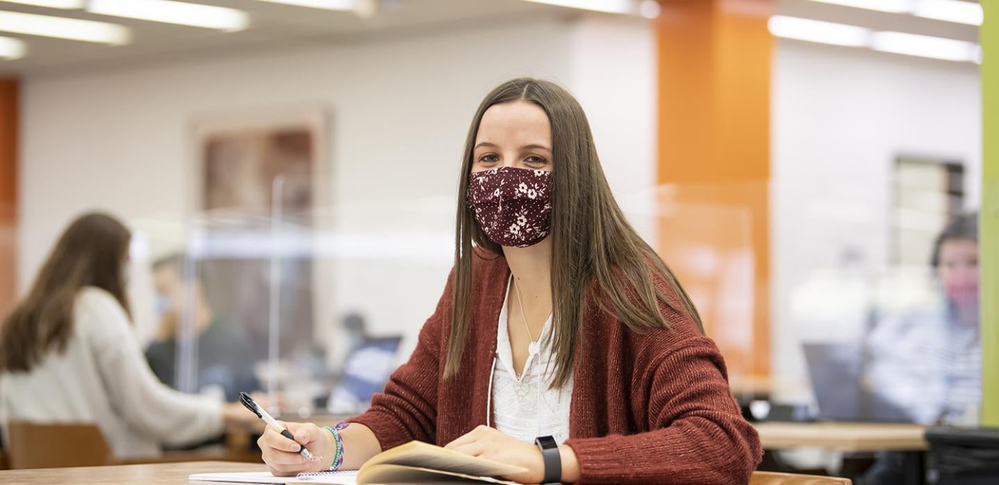 A student in an orange sweater and white top in a red face mask studying