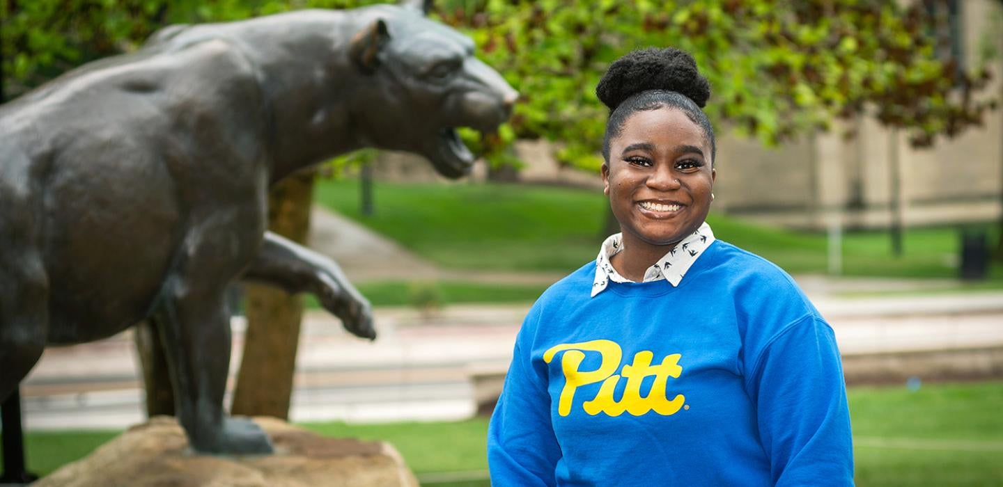 a woman in a blue and gold Pitt shirt smiling broadly in front of a Panther statue