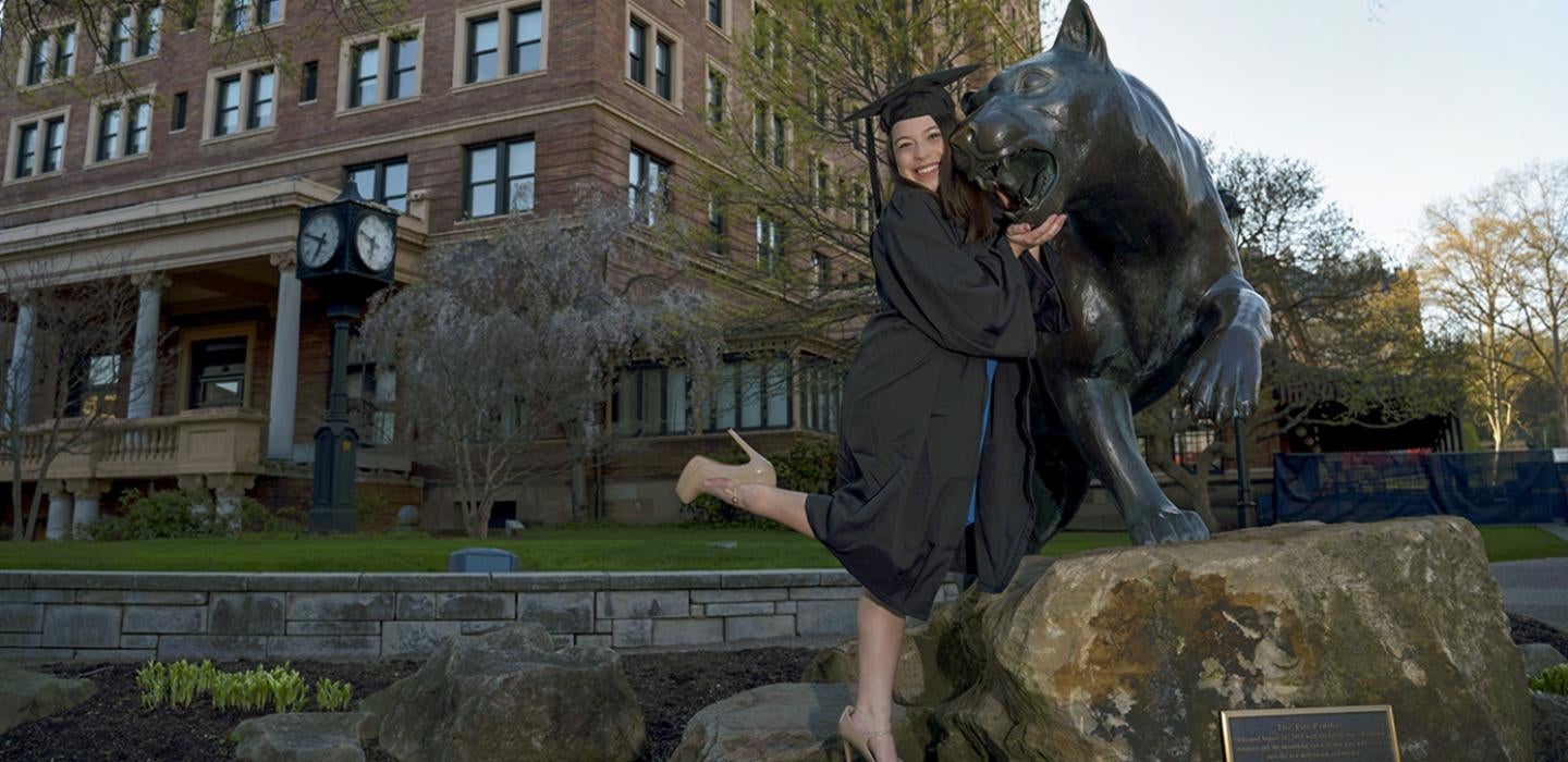 A woman in a black graduation cap and gown posing with a statue of a panther