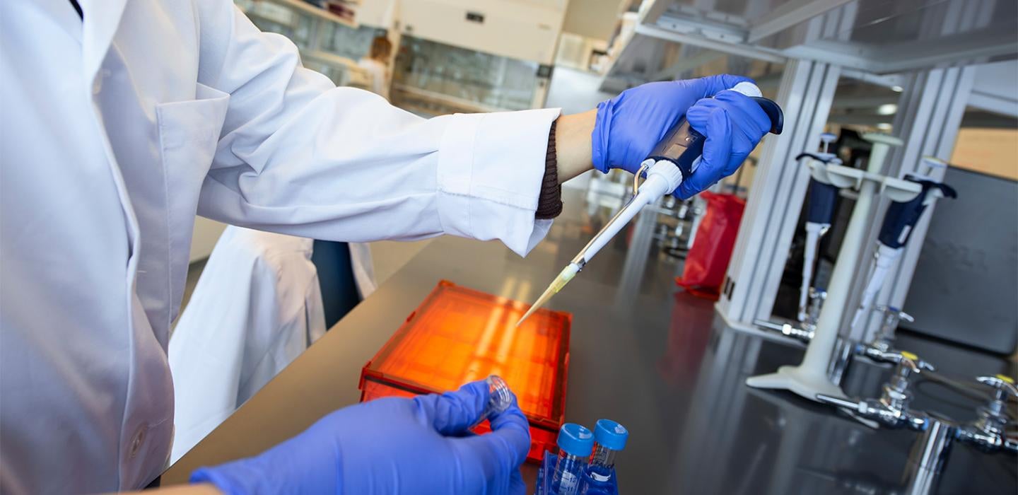A person in a white lab coat and blue gloves uses a pipette