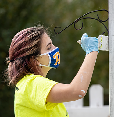 Woman in yellow shirt and Pitt mask painting white fence