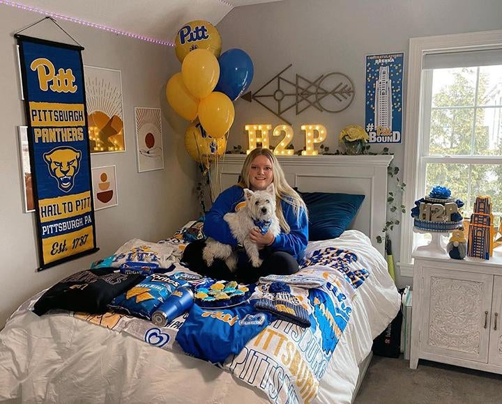 Girl and dog sitting on blue and yellow Pitt decorated bed 