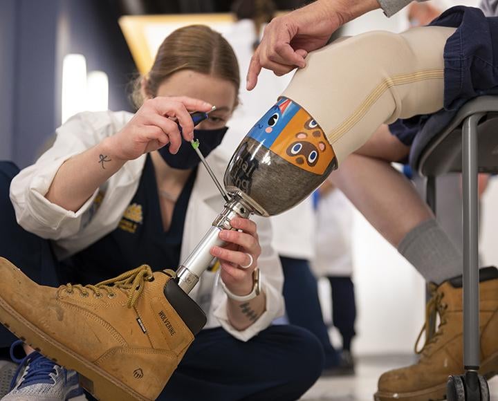 A student in a lab coat uses tools on a prosthetic leg.