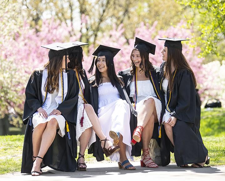 Five students wearing white dresses and black graduation robes sit on an outdoor bench smiling and laughing.