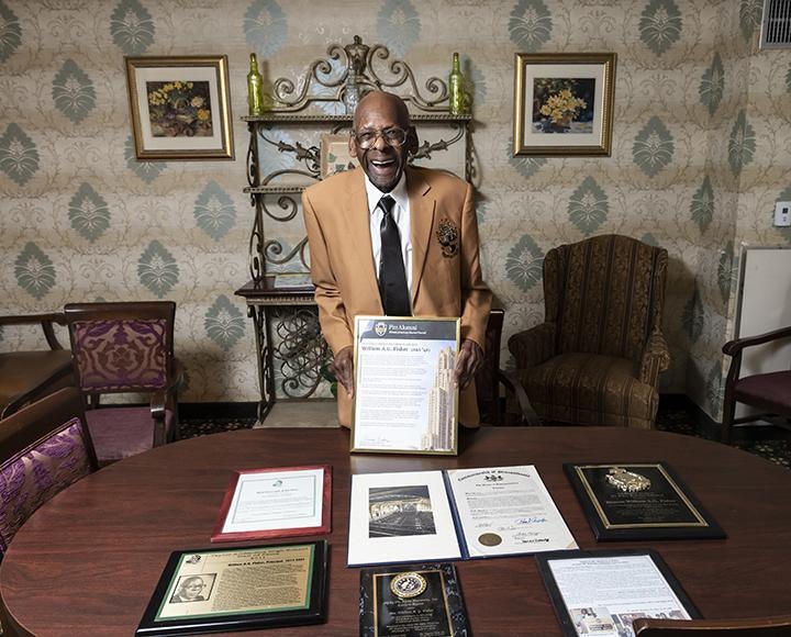 A man smiles while holding up a framed letter. Other awards and certificates sit in front of him on a wood table.