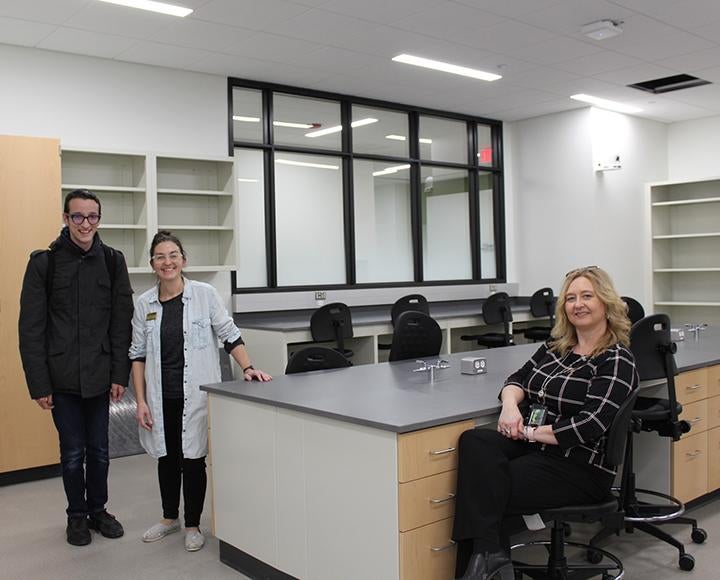 A person in a black jacket and a person in a white coat stand in a new lab. A person in a black shirt sits at one lab table