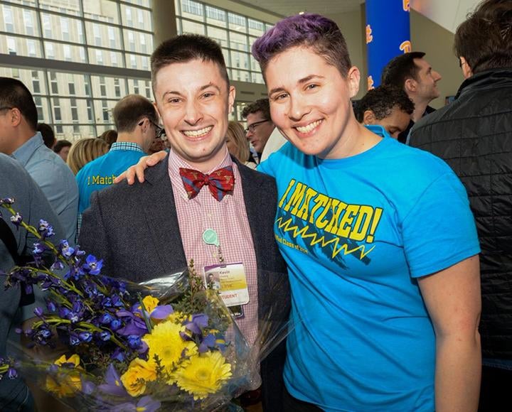 A person in a pink shirt and bow tie holds flowers and poses with a person in a blue match day T-shirt