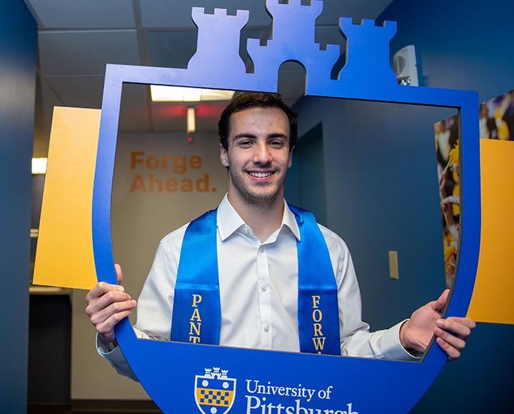 A person in a white shirt and blue stole poses with a University of Pittsburgh frame