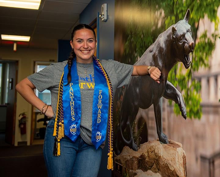 A person in braids and graduation cords poses with a Panther wall display