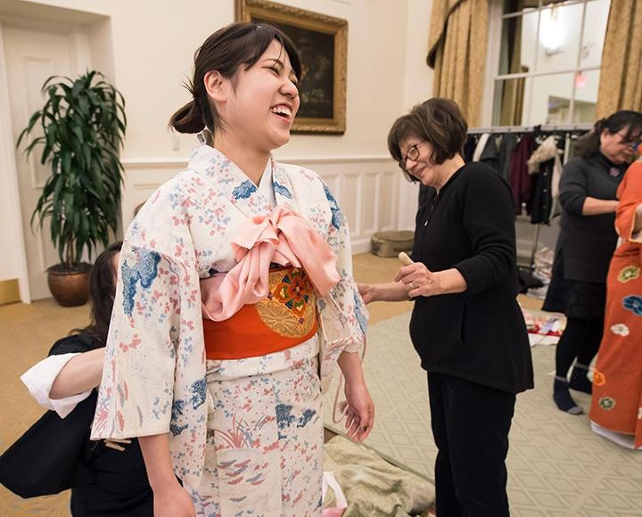 A girl in a white kimono with blue flowers laughs as a person adjusts a decorative orange belt behind her back