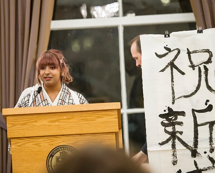 A girl in a white kimono speaks at a podium next to a painted Japanese phrase
