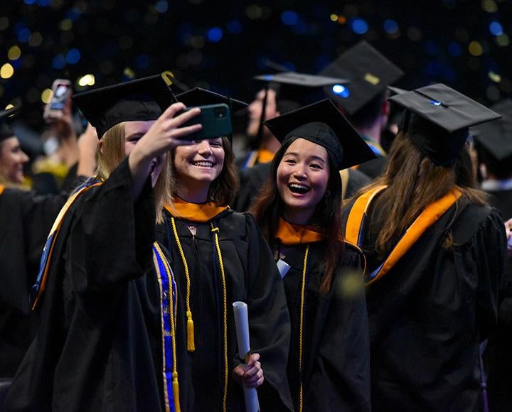 A person takes a selfie with two friends in graduation regalia