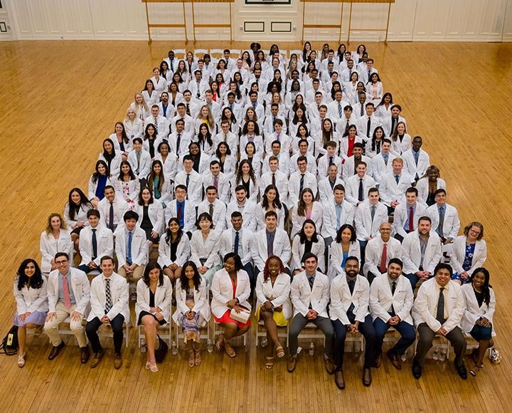 Students in white coats sit in rows of chairs for a photo taken from above