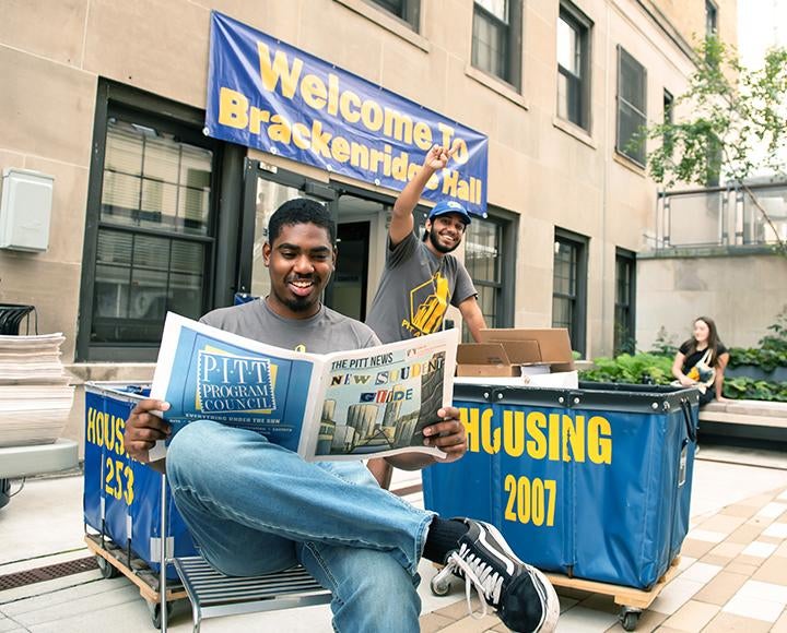 a student pushes a moving bin while another sits in the foreground reading a Pitt News paper