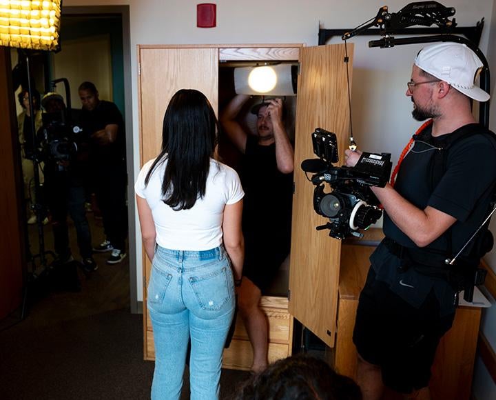 People film a person standing in a dorm room. A lighting operator stands in an open wardrobe.
