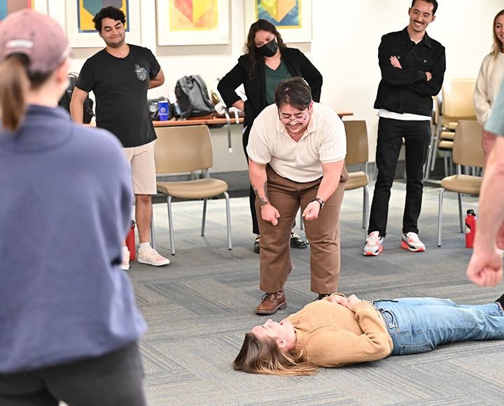 Everyone is laughing as one student stands over another who is laying on the ground in a classroom. Other students stand in a circle around them.