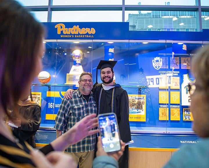 A graduate poses with his father in front of a Pitt basketball display