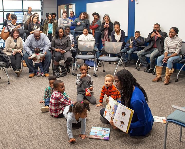 An adult reads a picture book to several toddlers on the floor as adults watch from a circle of chairs